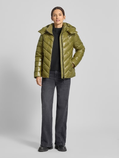 Modelka kobieta ma na sobie Steppjacke mit Kapuze und Reißverschluss od Christian Berg Woman Selection, kolor oliwkowy, krój regular fit, kaptur, zamek błyskawiczny, kieszenie z zatrzaskiem, długość tyłu w rozmiarze 36: 63 cm, szerokość ramion w rozmiarze 36: 42 cm, szerokość w klatce piersiowej w rozmiarze 36: 52 cm, długość rękawa w rozmiarze 36: 63 cm, widok całej sylwetki z przodu
