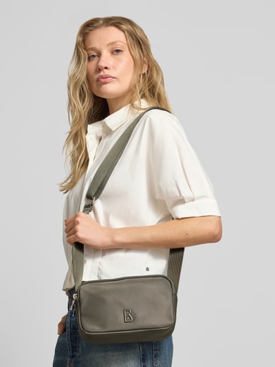 Frauenmodel trägt Handtasche mit Label-Detail Modell 'verbier play 1.0 avy' von BOGNER, Hauptfach mit Reißverschluss, verstellbarer Schulterriemen, Breite 22 cm, Länge 15 cm, Tiefe 7 cm, Länge Schulterriemen 116 cm, in neutralen Farbtönen, glatte Textur, Ganzkörperansicht von vorne.