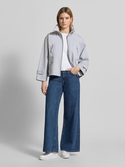 Modelka nosi kurtkę 'Jacke mit Stehkragen' od Blonde No. 8, bawełna, luźny krój, stójka, zamek błyskawiczny dwustronny, brustasza, wpuszczane kieszenie, długość tyłu w rozmiarze 36: 61 cm, szerokość ramion w rozmiarze 36: 51 cm, szerokość klatki piersiowej w rozmiarze 36: 60 cm, długość rękawów w rozmiarze 36: 60 cm, jasnoszara, gładka faktura, szerokie niebieskie jeansy, białe sneakersy, całopostaciowy widok z przodu.