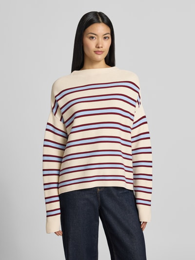 Frauenmodel trägt Loose Fit Pullover aus reiner Baumwolle Modell 'MERINAA' von ARMEDANGELS, creme mit roten und lilafarbenen Streifen, kombiniert mit dunkelblauer Jeans, Ganzkörperansicht von vorne.