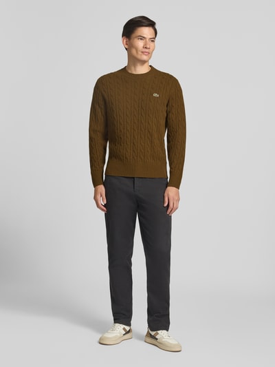 Männermodel trägt Regular Fit Pullover aus reiner Wolle von Lacoste in Braun, kombiniert mit dunkler Hose, Ganzkörperansicht von vorne.