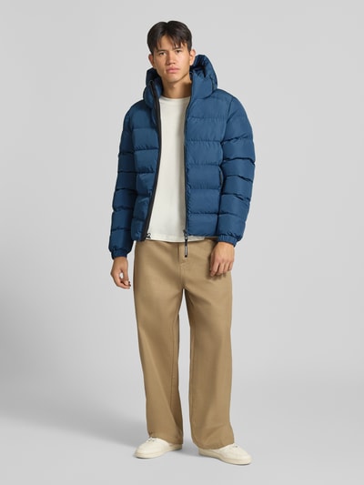 Männermodel trägt Steppjacke mit Label-Stitching und Kapuze von Superdry in Blau, kombiniert mit beiger Hose, Ganzkörperansicht von vorne. Herren Steppjacke, gerader Schnitt, Kapuze mit Kordelzug, Reißverschluss, seitliche Eingrifftaschen, elastische Ärmelabschlüsse, Rückenlänge 67 cm, Schulterbreite 48 cm, Brustweite 58 cm, Ärmellänge 72 cm.