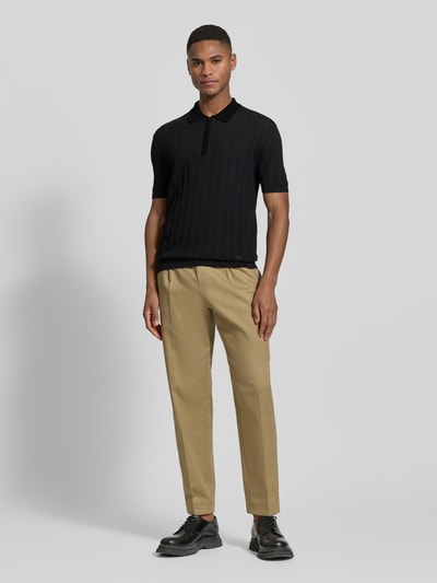 Männermodel trägt Body Fit Poloshirt mit kurzem Reißverschluss von Emporio Armani aus reiner Baumwolle mit gerippten Abschlüssen, Polokragen, in Schwarz, kombiniert mit beigefarbener Hose, Ganzkörperansicht von vorne.
