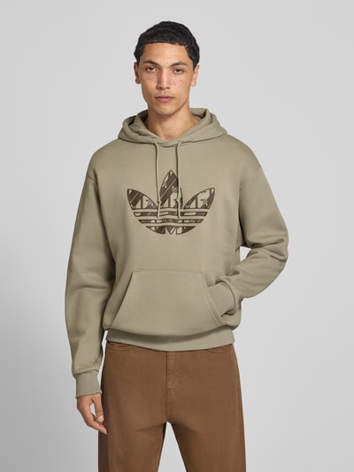 Männermodel trägt Regular Fit Hoodie aus Baumwoll-Mix mit Logo-Print von adidas Originals in Beige mit braunem Print, Nahaufnahme Oberkörper. Herren Hoodie von adidas Originals