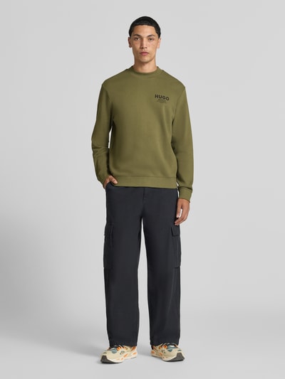 Męski model nosi Regular Fit Sweatshirt aus reiner Baumwolle model 'Nespon' von HUGO, oliwkowa zieleń, czarne spodnie, pełny widok z przodu, Herren Sweatshirt von HUGO Blue.
