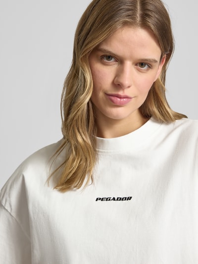 Kobiece model prezentuje damski t-shirt z bawełny Pegador Oversized Fit T-Shirt mit Label-Stitching model 'BRACY', okrągły dekolt, wyhaftowane logo, długość tyłu w rozmiarze S: 61 cm, jasny kolor, gładka faktura, zbliżenie na górną część ciała