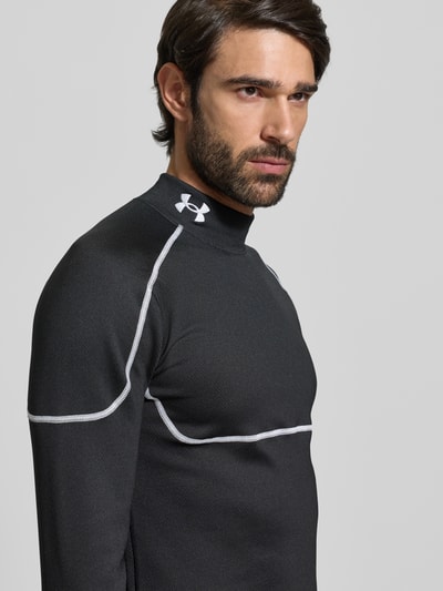 Männermodel trägt Funktionsshirt met opstaande kraag en label-stitching van Under Armour, zwart met witte naden, slim fit, materiaal-mix, detailopname bovenlichaam.