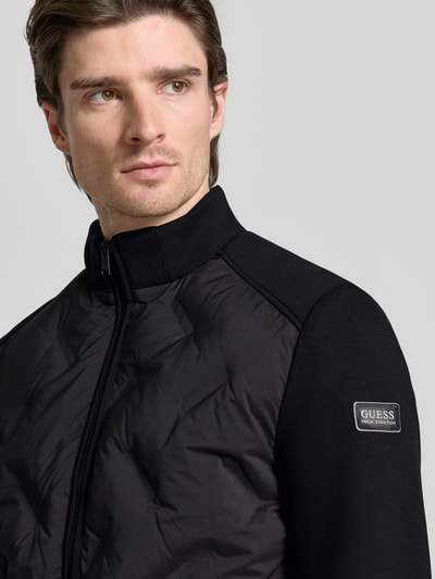 Männermodel trägt Regular Fit Jacke mit Stehkragen von Guess in Schwarz, Material-Mix, Reißverschluss, Nahaufnahme Oberkörper.