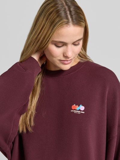 Frauenmodel trägt Oversized Fit Sweatshirt aus Baumwoll-Mix mit Stretch-Anteil von American Vintage in Dunkelrot mit Motiv-Print, Rundhalsausschnitt, elastische Bündchen, weiche Textur, Nahaufnahme Oberkörper.