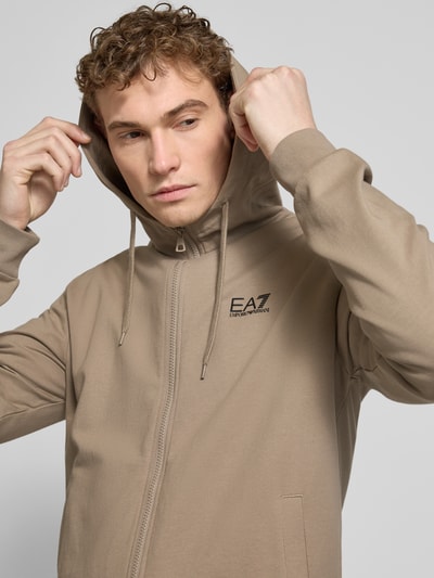 Męski model nosi Slim Fit Sweatjacke mit Kapuze od EA7 Emporio Armani, beżowy, bawełniany, logo, detal kaptura, zbliżenie na górę ciała.