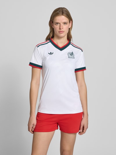 Kobiece model nosi Mexiko Auswärtstrikot 2026 od adidas Sportswear, efekt: wielokolorowy, krój regular fit, faktura: fakturowany wzór, aplikacja z logo, wyhaftowane logo, paski w kontrastowym kolorze, dekolt w serek, krótki rękaw, zakończenie rękawów: prążkowane, lamówki, szybkoschnący, materiał regulujący wilgoć, całkowity widok od przodu.