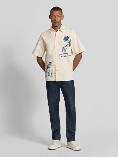 Männermodel trägt Relaxed Fit Freizeithemd aus reiner Baumwolle von Scotch & Soda, creme mit Strukturmuster, Blau-Motiv-Stitching, Ganzkörperansicht von vorne.