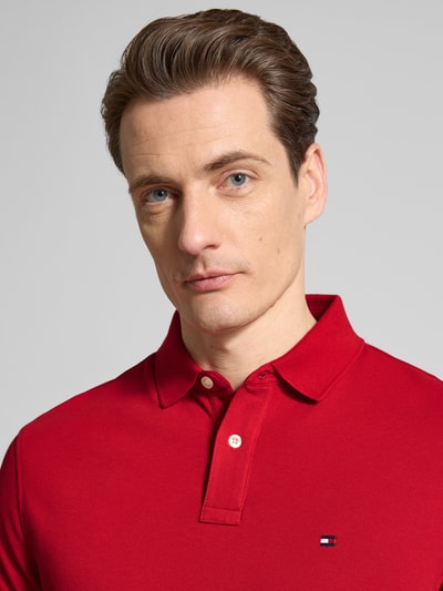 Mężczyzna nosi Regular Fit Poloshirt aus Baumwoll-Mix von Tommy Hilfiger w kolorze czerwonym, logo z przodu, materiał pique, bawełniany mix, krótki rękaw, krój regular fit, kołnierzyk polo, zapięcie na dwa guziki, widoczne przeszycia, jednolity kolor, aplikacja logo, ujęcie: zbliżenie na górną część ciała
