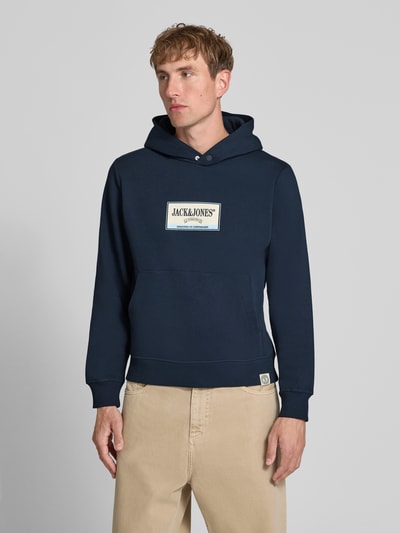 Männermodel trägt Hoodie mit Label-Print Modell 'FREDERIKSBERG' von JACK & JONES, dunkelblau, Kängurutasche, Ganzkörperansicht von vorne. Baumwoll-Mix, lockerer Schnitt, gerippte Abschlüsse, Kapuze, Rückenlänge 65 cm, Ärmellänge 67 cm.