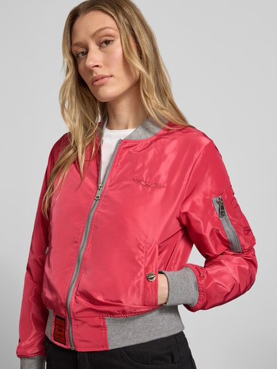 Frauenmodel trägt Bomberjacke mit Label-Patch und Reißverschlusstasche am Ärmel von Original Bombers in Rot mit grauen Abschlüssen, Ganzkörperansicht von vorne, Lockerer-Schnitt, Stehkragen, Reißverschluss, Logo-Stitching, Eingrifftaschen mit Druckknopfverschluss, Rückenlänge bei Größe S: 53 cm, Schulterbreite: 37 cm, Brustweite: 48 cm, Ärmellänge: 63 cm.