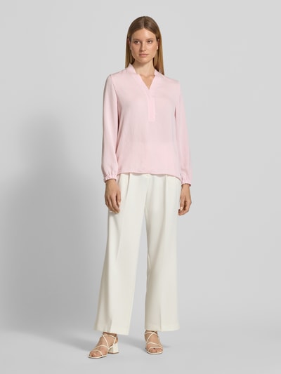 Kobiece model nosi Regular Fit Bluse mit V-Ausschnitt von Marc Cain, krój regular fit, dekolt w serek, detal z logo, długość tyłu w rozmiarze 36: 65 cm, długość rękawów w rozmiarze 36: 59 cm, jasnoróżowa bluzka, jasne szerokie spodnie, jasne sandały, gładkie tkaniny, zdjęcie w pełnej sylwetce z przodu.