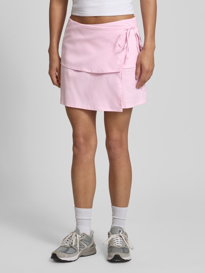 Frauenmodel trägt Minirock mit Leinen-Anteil Modell 'Sina' von JJXX in Rosa, Zierschleife, Wickel-Optik, leichte A-Linie, Nahaufnahme Oberkörper. Bundweite bei Größe S: 38 cm, elastischer Baumwoll-Mix mit Leinen-Anteil.