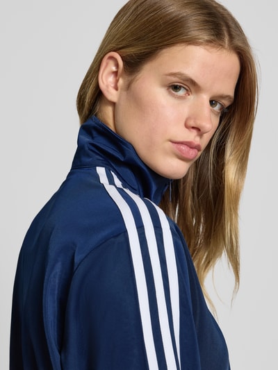 Vrouwenmodel draagt Sweatjacke mit Stehkragen van adidas Originals in donkerblauw met witte strepen, label-stitching, detailopname.