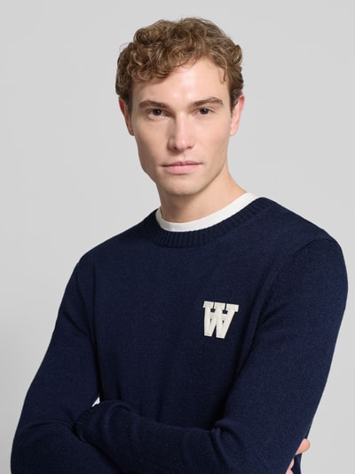 Männermodel trägt Strickpullover aus Lammwolle Modell 'Tay' von Wood Wood in Dunkelblau mit Label-Stitching, Rundhalsausschnitt, Regular-Fit, Nahaufnahme Oberkörper. Rückenlänge bei Größe M 72 cm, Ärmellänge 68 cm.