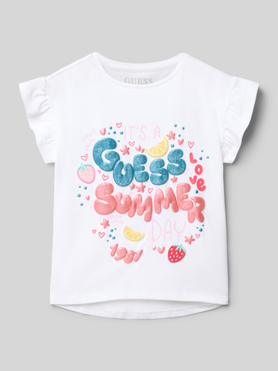 Mädchen trägt Regular Fit T-Shirt mit Statement-Print von Guess aus elastischem Baumwoll-Mix, Weiß mit buntem Motiv- und Logo-Print, Rüschenbesatz, Rundhalsausschnitt, Rückenlänge 44 cm bei Größe 116, Ganzkörperansicht von vorne.