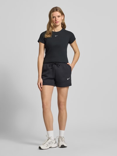 Frauenmodel trägt Cropped T-Shirt in Ripp-Optik mit Logo von Nike, Schwarz, Baumwoll-Elasthan-Mix, Rundhalsausschnitt, Rückenlänge 48 cm, Ganzkörperansicht von vorne.