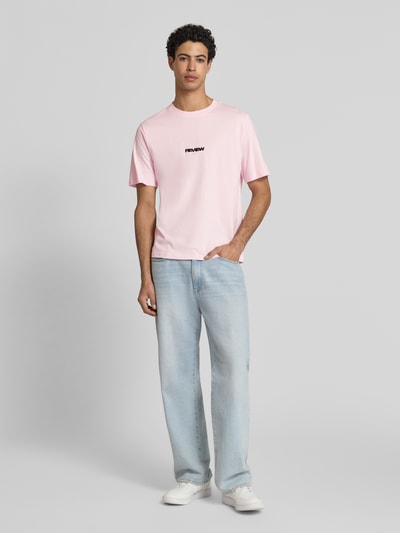 Mężczyzna nosi T-Shirt mit Logo und Rundhalsausschnitt od REVIEW, bawełna, krój relaxed fit, długość tyłu 68 cm, jasnoróżowy; całopostaciowy widok z przodu.