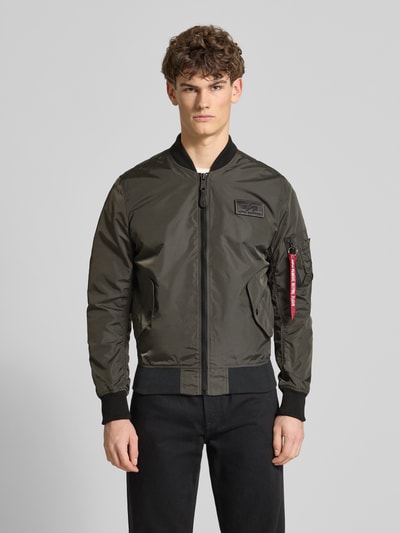 Męski model nosi Regular Fit Bomberjacke mit Logo-Patch od Alpha Industries w kolorze oliwkowym, stójka, łatka z logo, kieszenie, zamek, kieszeń na rękawie, długość tyłu w rozmiarze M: 65 cm, szerokość ramion w rozmiarze M: 45 cm, szerokość klatki piersiowej w rozmiarze M: 54 cm, długość rękawa w rozmiarze M: 69 cm, widok całej sylwetki z przodu.