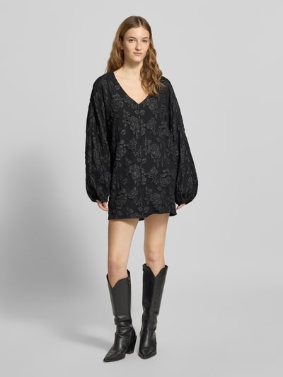 Vrouwenmodel draagt Minikleid mit V-Ausschnitt van Gina Tricot, los vallend, V-hals, sierbies, elastische mouwuiteinden, zwart met bloemenpatroon, volledige lichaamsweergave van voren