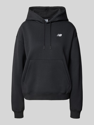 Frauenmodel trägt Regular Fit Hoodie aus Baumwoll-Mix von New Balance in Schwarz mit Logo-Print, Känguru-Tasche, Kapuze, Ganzkörperansicht von vorne. Damen Hoodie aus Baumwoll-Mix, Regular Fit, elastische Abschlüsse, Tunnelzug, Innenbeinlänge S: 60 cm.