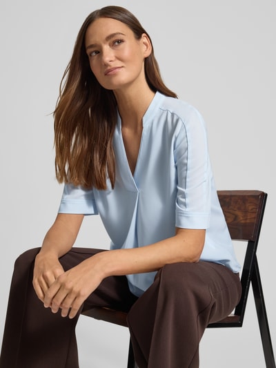 Modelka nosi Regular Fit Bluse mit V-Ausschnitt od Marc Cain, jasnoniebieska, stójka, krój regular fit, długość tyłu 59 cm, długość rękawa 29 cm, zdjęcie w zbliżeniu na górną część ciała.