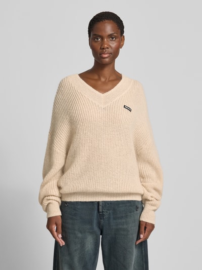 Frauenmodel trägt Oversized Pullover mit Woll-Anteil von ROTATE im Material-Mix, Strick-Optik, V-Ausschnitt, Logo-Patch, beige, Nahaufnahme Oberkörper.