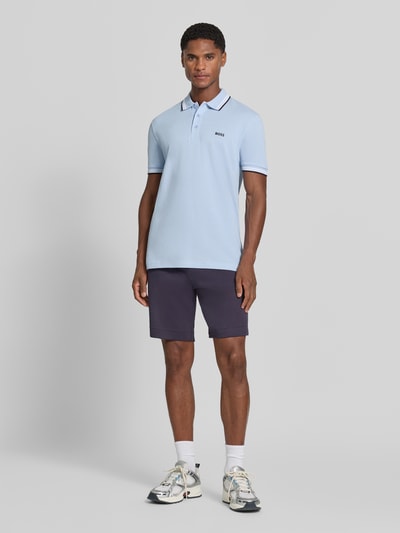Männermodel trägt Regular Fit Poloshirt mit Label-Stitching Modell 'Paddy' von BOSS in Hellblau, Seitentaschen-Shorts, Ganzkörperansicht von vorne. Reine Baumwolle, Polokragen, kurze Knopfleiste, gerippte Ärmelabschlüsse, Rückenlänge bei Größe M 73 cm.
