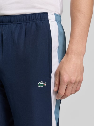 Männermodel trägt Regular Fit Trainingshose mit verstellbarem Gummibund von Lacoste in Dunkelblau mit weißen und blauen Streifen, Nahaufnahme Oberkörper, Herren Trainingshose von Lacoste Sport.