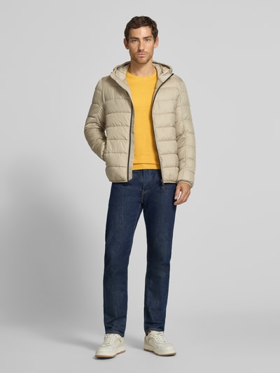 Männermodel trägt Strickpullover mit geripptem Rundhalsausschnitt von MCNEAL in Gelb, blaue Jeans, beige Steppjacke, Ganzkörperansicht von vorne. Taillierte Passform, leicht überschnittene Schultern, gerippte Abschlüsse, Rückenlänge 70 cm, Ärmellänge 65 cm.