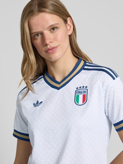 Frauenmodel trägt Italien Auswärtstrikot 2026 von adidas Sportswear, mehrfarbig, Regular Fit, V-Ausschnitt, Logo-Stitching, Kontraststreifen, kurzarm, Nahaufnahme Oberkörper.