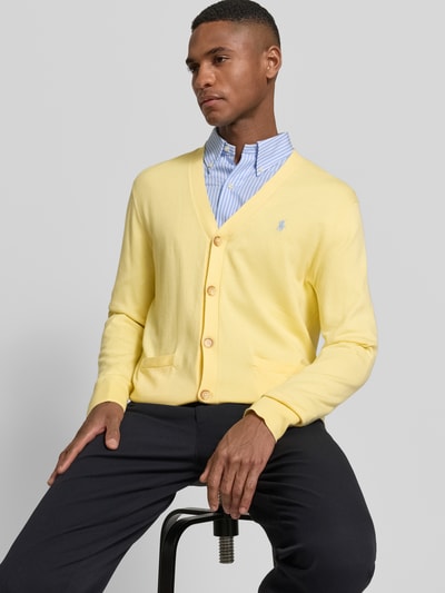 Männermodel trägt Regular Fit Gebreid jack uit puur katoen van Polo Ralph Lauren in effen geel met logodetail, knoopsluiting, opgestikte zijzakken, geribd aan mouwboord en rompzoom, diepe V-hals en ronde hals, knitweartextuur, voorkant ganzkörperansicht.