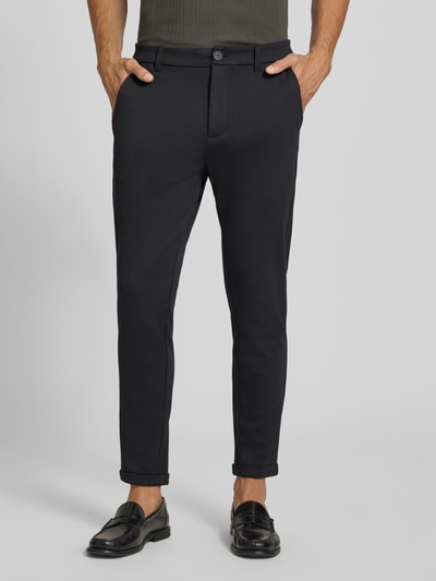Męski model nosi Slim Fit Hose mit Viskose-Anteil in verkürzter Passform von Lindbergh, czarny, mieszanka wiskozy, wpuszczane i tylne kieszenie, zapięcie na guzik i zamek błyskawiczny, szerokość w rozmiarze M: 42 cm, długość wewnętrzna nogawki: 72 cm, krój slim fit, skrócony krój, materiał o gładkiej fakturze, spodnie materiałowe, Ganzkörperansicht z przodu.