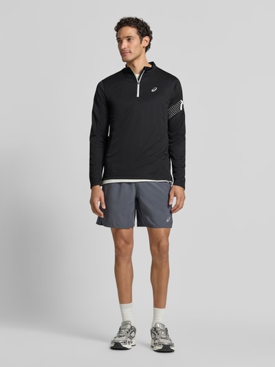 Męskimodel nosi bluzkę z długim rękawem Model 'ICON' od ASICS TIGER w kolorze czarnym z nadrukiem z logo, prosty krój, kołnierz zapinany, długość tyłu w rozmiarze M: 72 cm, zdjęcie całej sylwetki z przodu.
