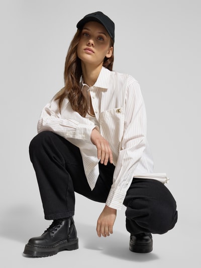 Kobiece model prezentuje Relaxed Fit Bluse mit Brusttasche und Label-Stitching von Calvin Klein Jeans, biała bawełna, kieszeń na piersi, krój relaxed fit, kołnierzyk koszulowy, subtelne paski, Szerokość w rozmiarze S 69 cm, długość rękawa 58 cm, widok całej sylwetki z przodu.