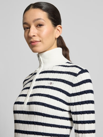 Frauenmodel trägt Strickpullover mit Stehkragen und Reißverschluss von Gant in Weiß mit dunkelblauen Streifen, Ripp-Optik, Nahaufnahme Oberkörper. Baumwoll-Mix mit Stretch-Anteil, Slim Fit, Label-Stitching.