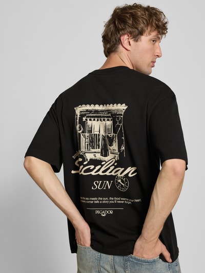 Mannenmodel draagt zwart oversized t-shirt van puur katoen, logoprint en motiefprint op de achterkant, Model 'Pedroche' van Pegador, ruglengte maat M 71 cm, ganzlich achteraanzicht.