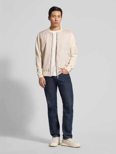 Männermodel trägt Collegejacke mit Zweiwege-Reißverschluss von MAERZ Muenchen aus puur katoen, regular fit, opstaande kraag, steekzakken, jeans en lichte sneakers, neutrale tinten, gladde textuur. Ganzkörperansicht von vorne.