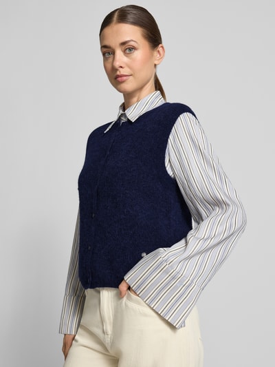Frauenmodel trägt Regular Fit Cardigan aus elastischem Alpaka-Mix mit Knopfleiste von MOS MOSH, ärmellos, dunkelblau, glatte Textur, kombiniert mit cremefarbenen Hosen und gestreifter Bluse, Nahaufnahme Oberkörper.