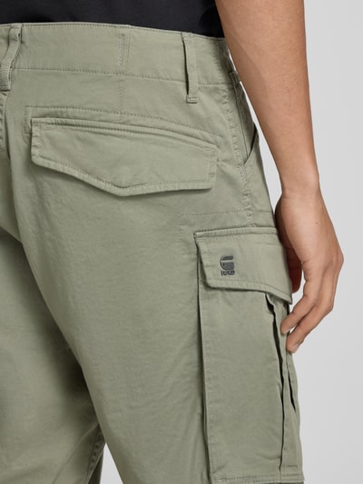 Männermodel trägt bermudy z mieszanki bawełny z dodatkiem streczu G-Star Raw, model 'ROVIC ZIP', kieszenie z patką, zamek błyskawiczny, 5 kieszeni, szerokość w rozmiarze 32: 44 cm, długość wewnętrzna nogawki w rozmiarze 32: 28 cm, detal z logo, matowa tekstura, zielony kolor, detailaufnahme.