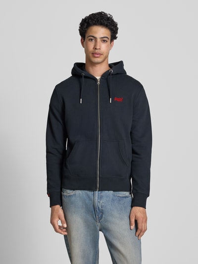 Männermodel trägt Sweatjacke mit Kapuze und Logo-Stitching von Superdry aus Baumwoll-Mix, Regular-Fit, Ganzkörperansicht von vorne, seitliche Eingrifftaschen, Reißverschluss, gerippte Abschlüsse, Label-Detail, Rückenlänge bei Größe M 66 cm, Ärmellänge bei Größe M 62 cm, dunkle Farbe, glatte Textur.