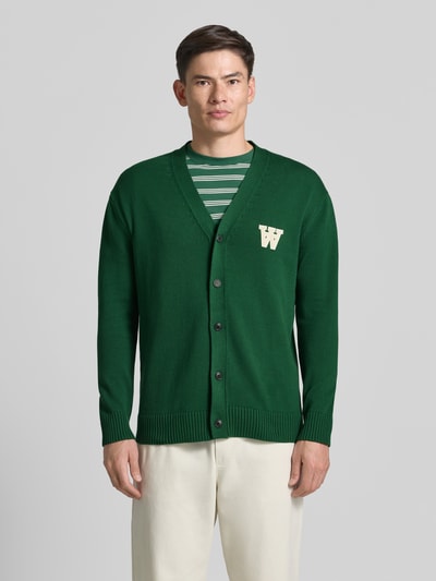 Männermodel trägt grüne Strickjacke mit V-Ausschnitt Modell 'Tay' von Wood Wood aus Reine Baumwolle, Gerader-Schnitt, Ganzkörperansicht von vorne.