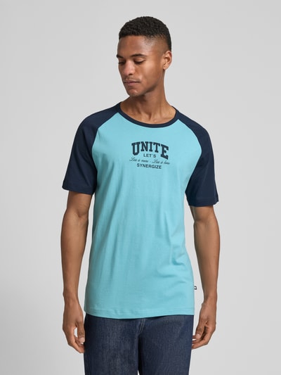 Męski model nosi Regular Fit T-Shirt aus reiner Baumwolle von s.Oliver RED LABEL, niebieskie dwa odcienie, nadruk statement, krótkie rękawy, widok od przodu, ujęcie do pasa.