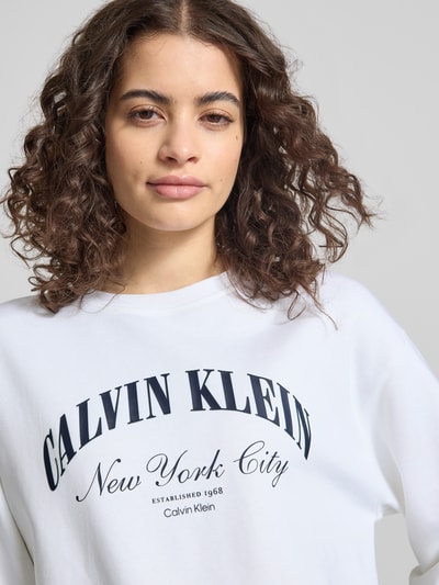 Kobiece modelka nosi bluzę Sweatshirt mit Label-Print Modell 'GRAPHIC' od Calvin Klein Jeans z mieszanki bawełny i elastanu, luźny krój, nadruk z logo, okrągły dekolt, białe tło, gładka tekstura, detale czarnego nadruku, zbliżenie górnej części ciała.