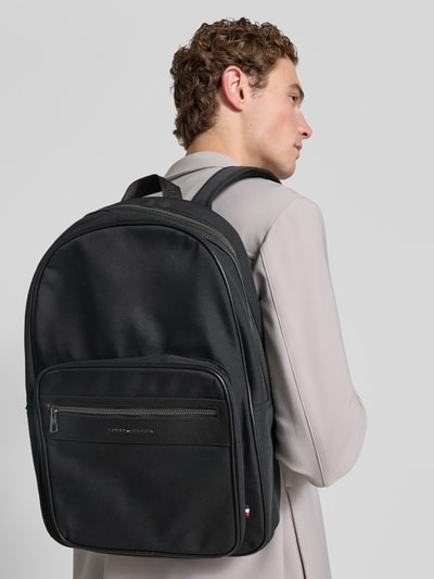 Männermodel trägt Rucksack mit Label-Schriftzug Modell 'REPREVE' von Tommy Hilfiger, Schwarz, Optik: Unifarben, Ganzkörperansicht Rückansicht, Herren Rucksack mit Reißverschluss, Aufteilung: Seitentasche, Hauptfach, Reißverschlussfach, Einschubfächer, Applikation: Label-Schriftzug, Trageoptionen: Tragehenkel, Schulterriemen, Materialverarbeitung: Webware.