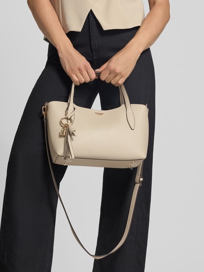 Frauenmodel trägt Umhängetasche mit Label-Applikation von GUESS in Beige mit Magnetverschluss, Clutch und Tragehenkel, Ganzkörperansicht von vorne.